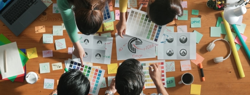 Illustration of graphic designers collaborating at a table, surrounded by laptops, sketchpads, and design tools, working together on creative projects.